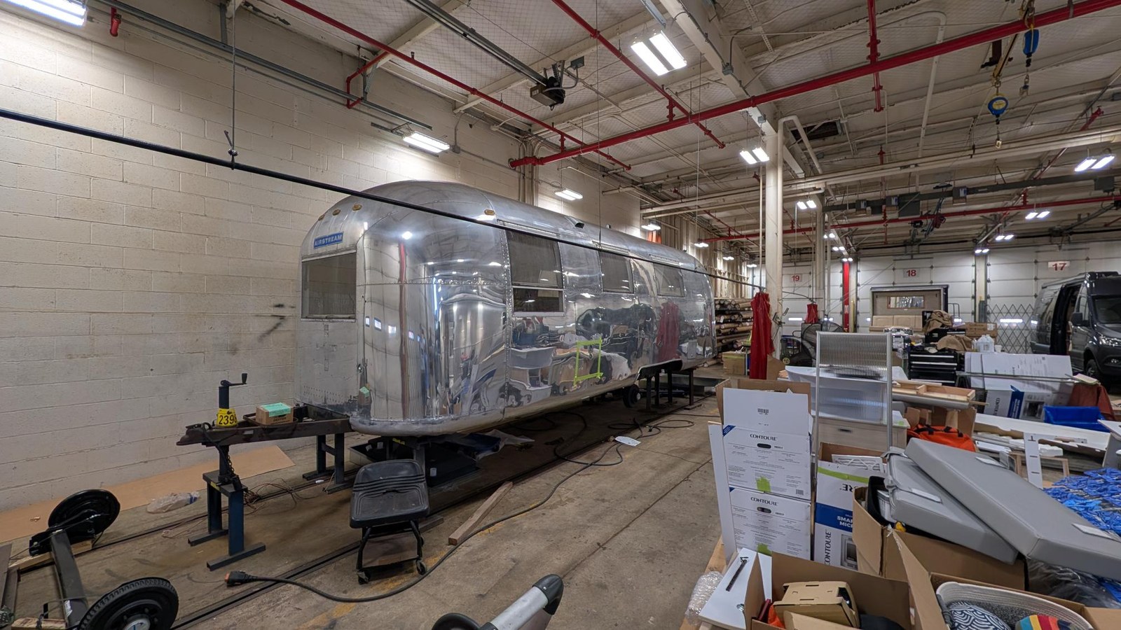 The 1967 Airstream Overlander up on shop stands inside a service bay at the Airstream Factory Service Center, its polished aluminum shell reflecting the length of the shop.